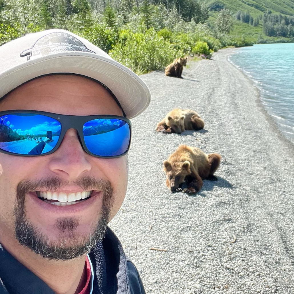 Man with round face, wearing wrap sunglasses and smiling in front of bears on the beach