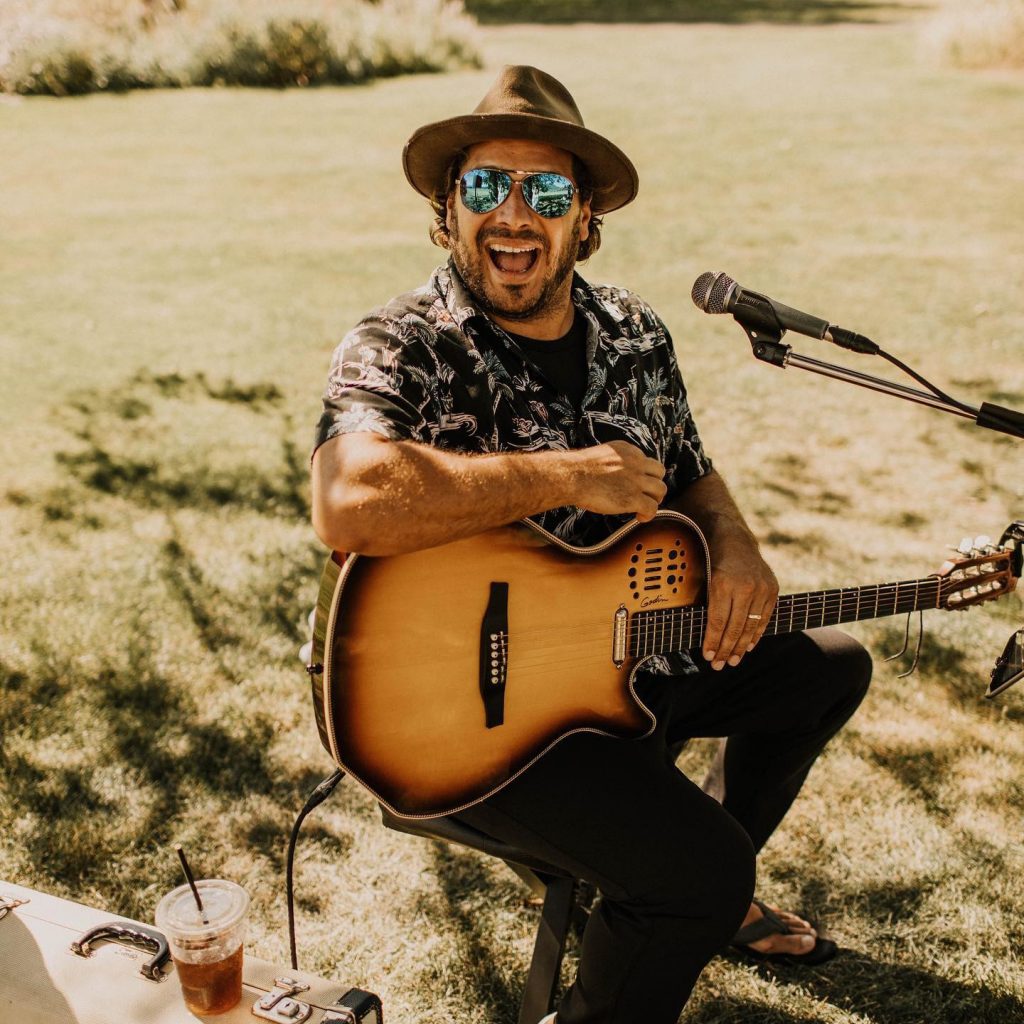 A man in blue shades sits on a lawn playing the guitar
