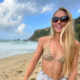 Woman in Maui Jim Starfish Sunglasses poses in swimwear in front of a scenic beach.