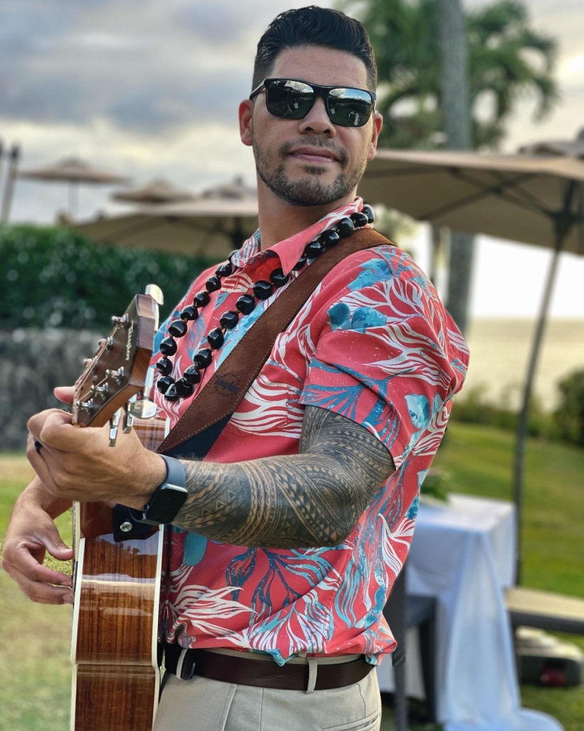 Man in Hawaiin shirt and black sunglasses plays a guitar