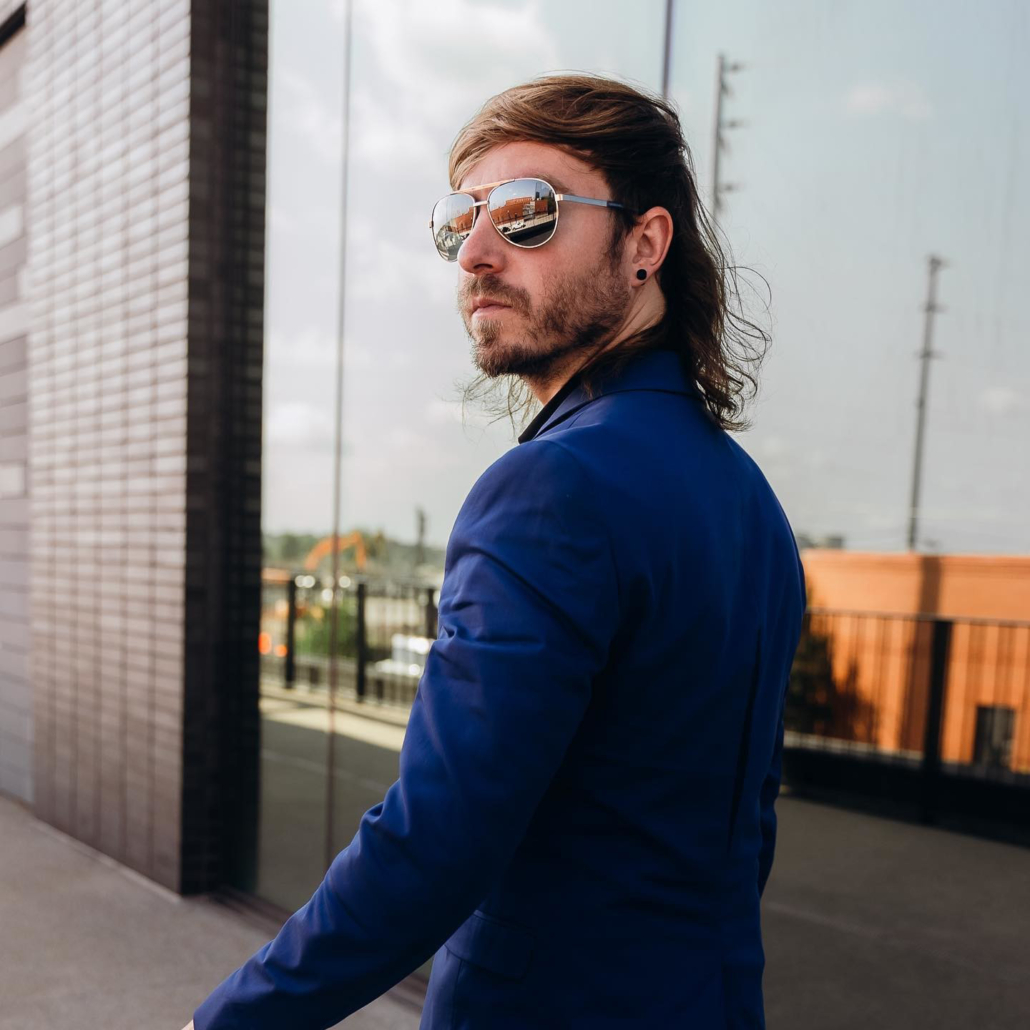 Man in blue suit and aviator sunglasses poses in front of a building